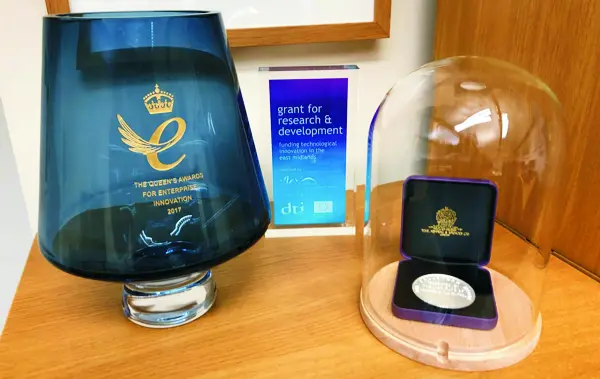 Three awards on a shelf: a blue glass Queen's Award for Enterprise trophy marked Innovation 2017, a small blue DTI grant for research and development award, and the Callendar Medal in its case under a glass dome.