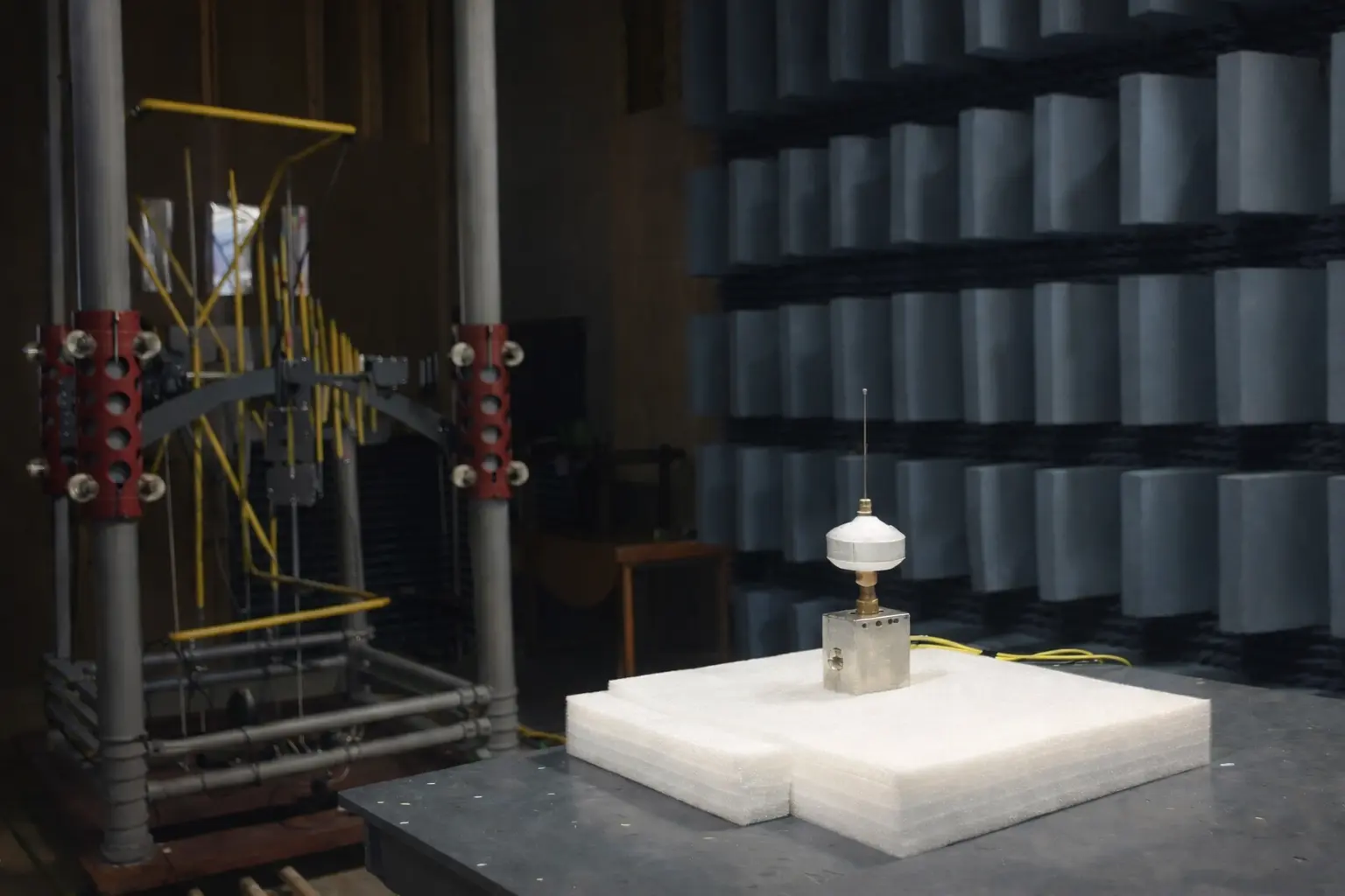 The Johnson noise thermometer's sensor head, its lid open, mounted on a block of RF-absorbing foam in an anechoic chamber, with a biconical and log-periodic antenna array on a mast to one side and rows of pyramidal foam absorber lining the chamber walls behind it.