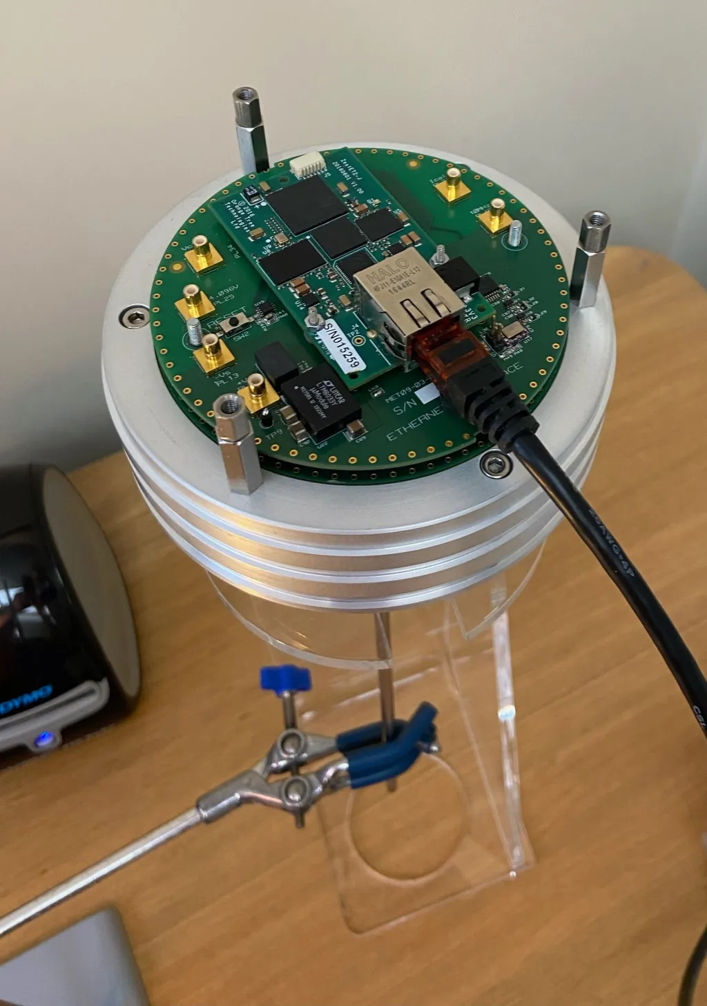 The Johnson noise thermometer with its lid off, showing a circular circuit board carrying the processing and Ethernet interface electronics, SMA connectors around the edge, mounted above the finned enclosure on a clear stand.