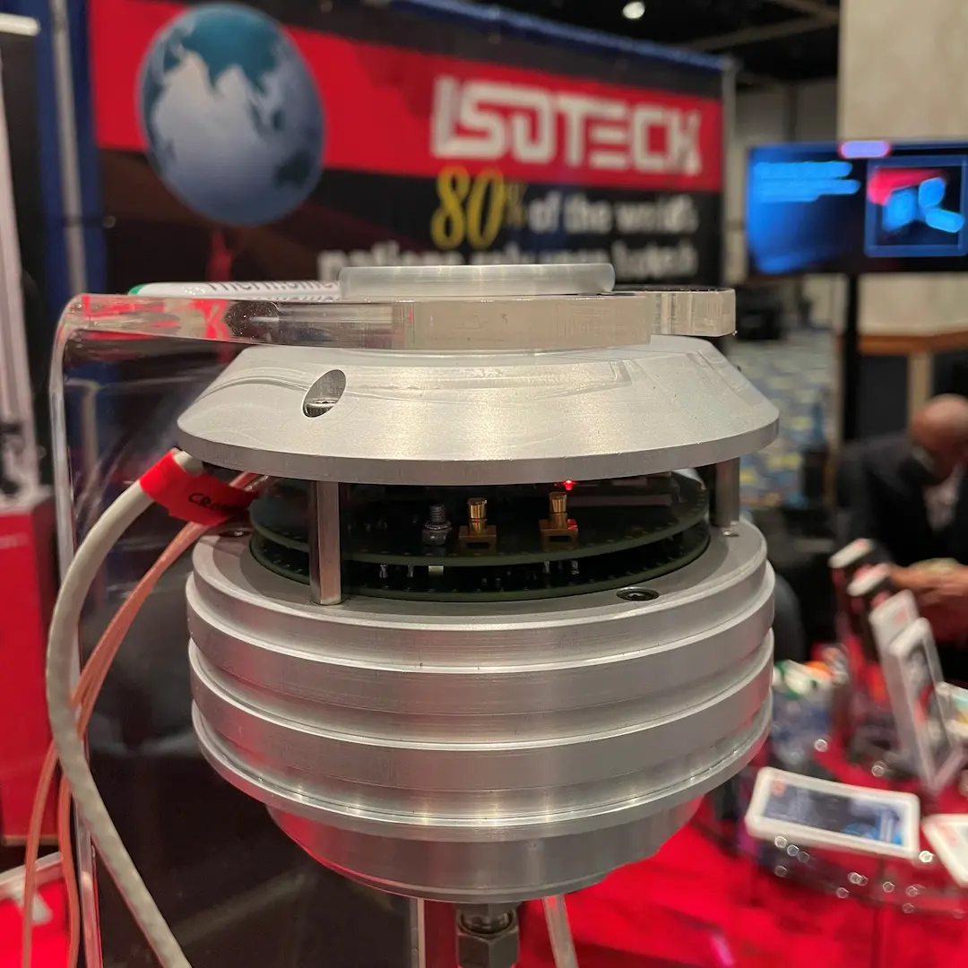 The Johnson noise thermometer’s sensor head on its clear acrylic stand at a trade-show booth, its aluminium lid lifted to show the internal circuit board with two brass connectors and a lit red indicator, an Isotech banner reading ‘80% of the world’s nations use Isotech’ visible behind it.