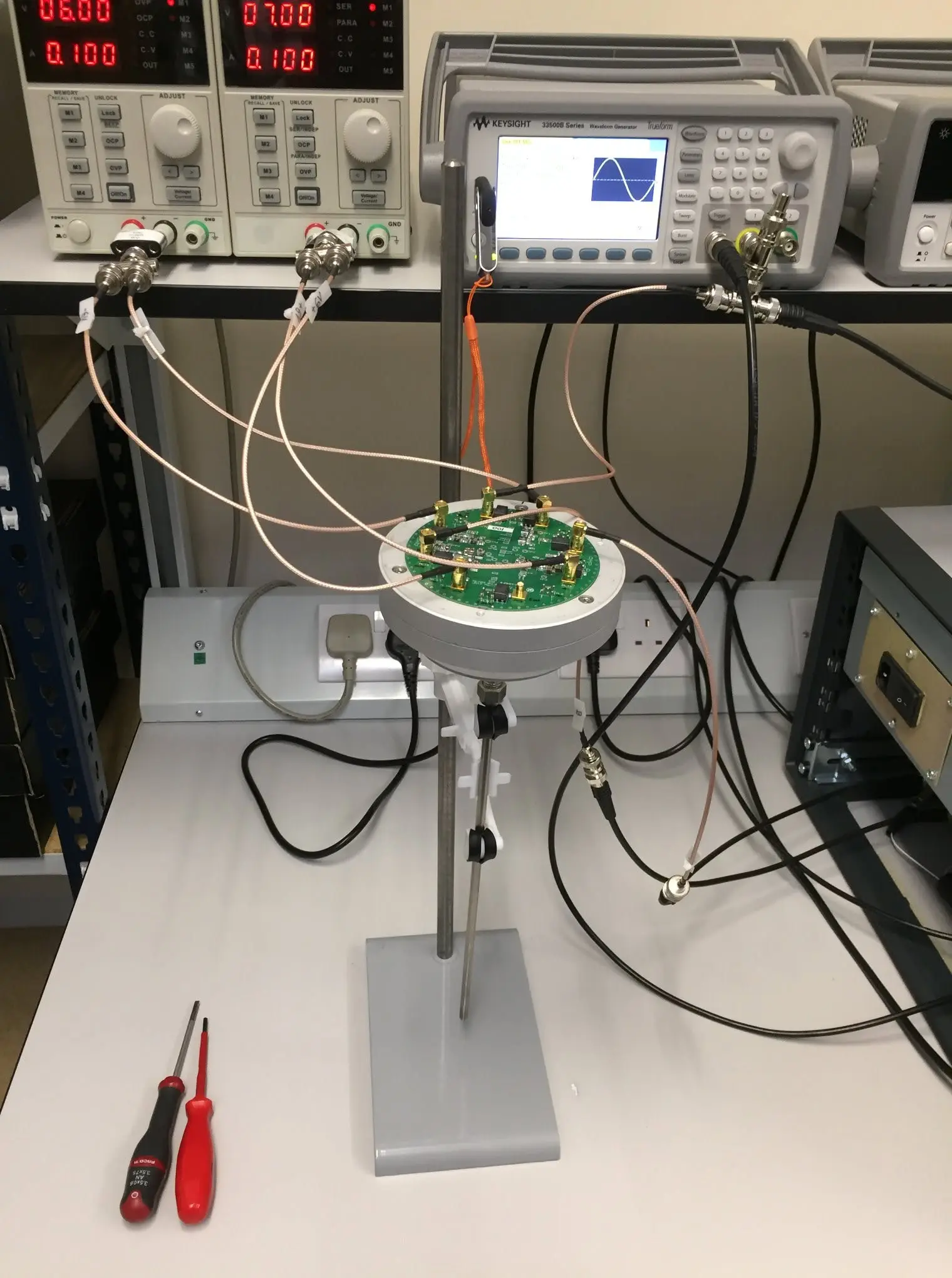 The JNT2 pre-amplifier board mounted on a stand on a lab bench, wired with SMA test cables to bench power supplies and a Keysight waveform generator during characterisation.
