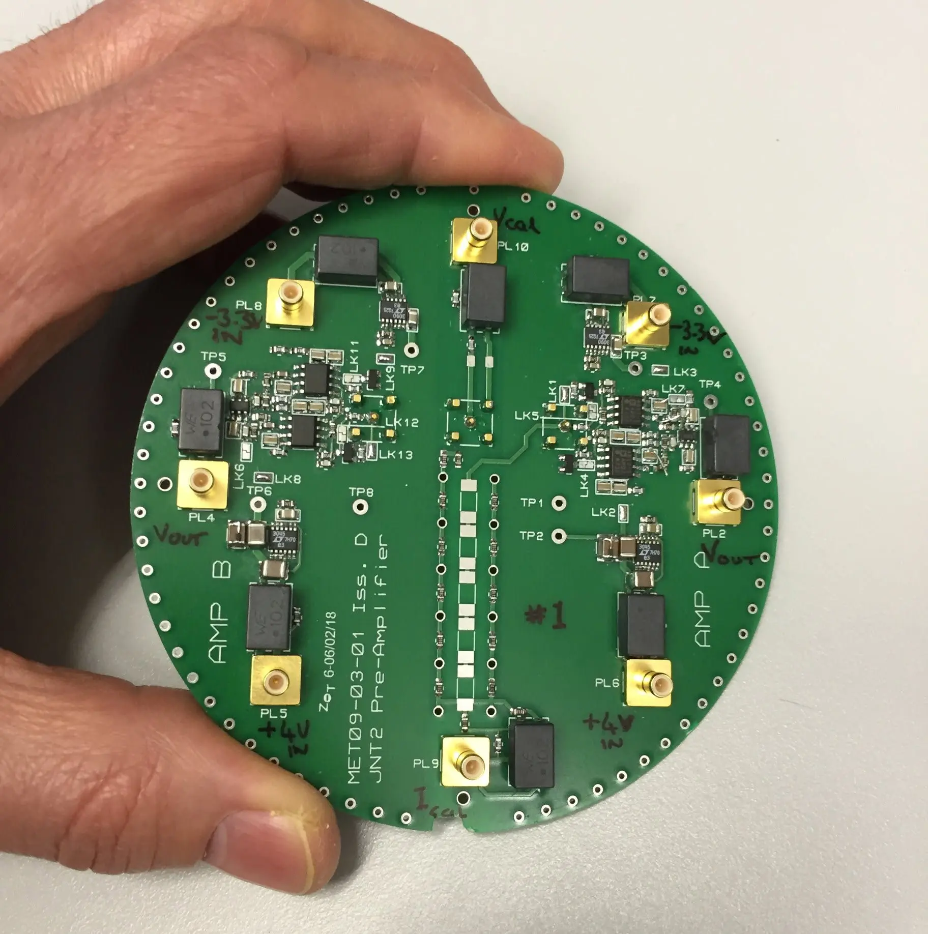 A circular circuit board labelled JNT2 Pre-Amplifier, held in one hand, with two mirrored amplifier sections marked AMP A and AMP B, SMA connectors around the edge and hand-written test-point labels.