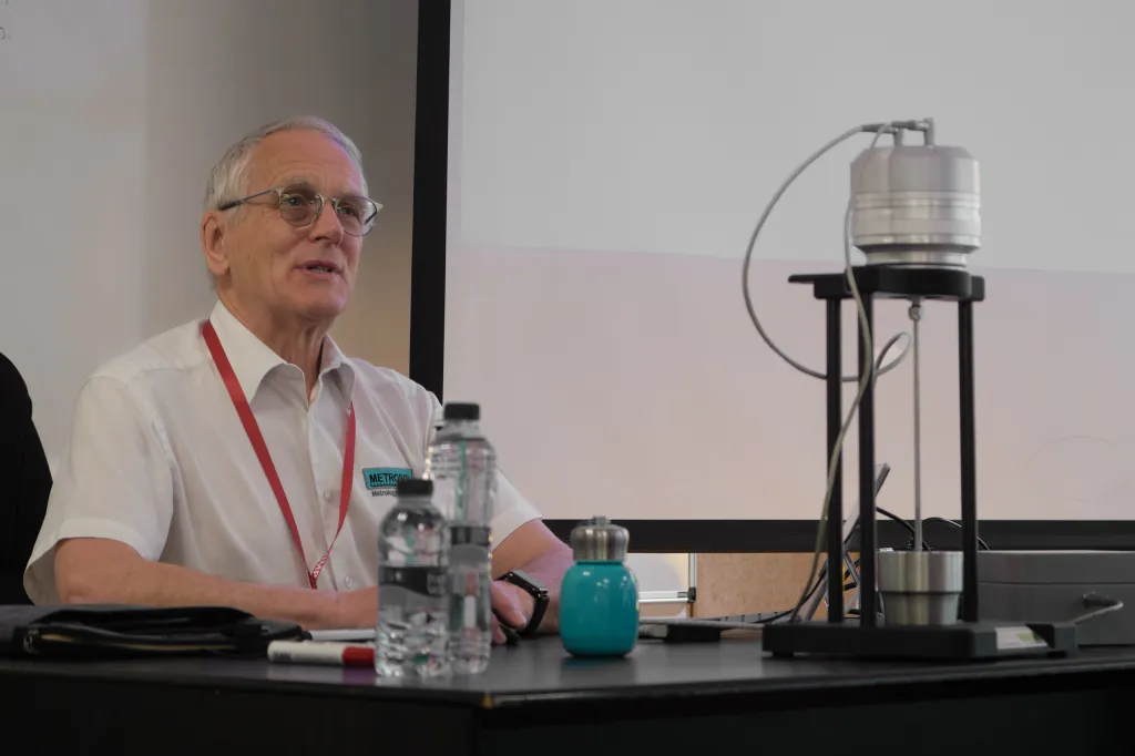 Paul Bramley speaking at a table, with the Johnson noise thermometer prototype set up on a stand beside him, its probe running down into a metal block.