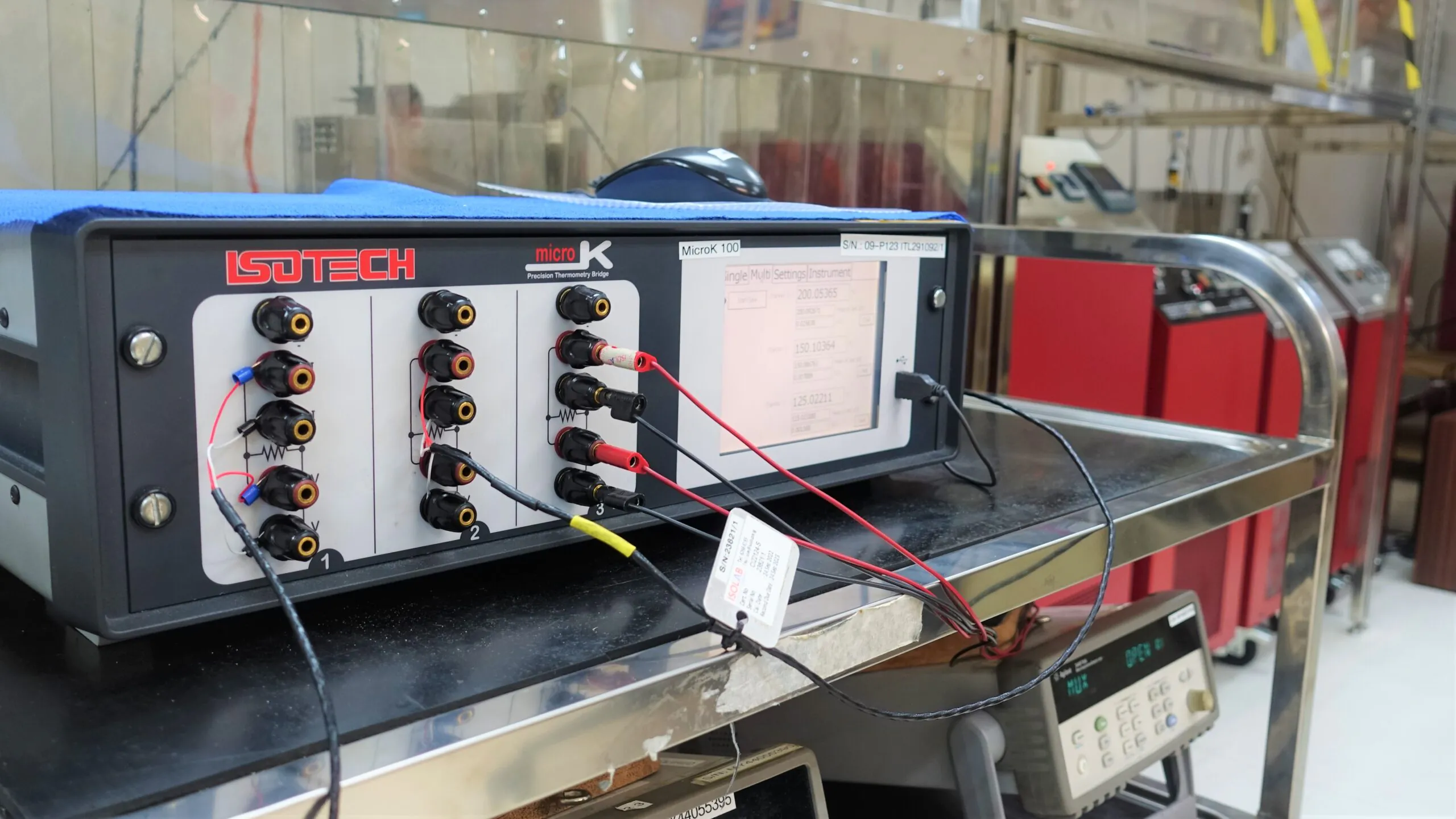 A microK 100 on a laboratory bench with red and black measurement leads running from its terminals, a calibration tag hanging from the cabling, and other instruments in the background.