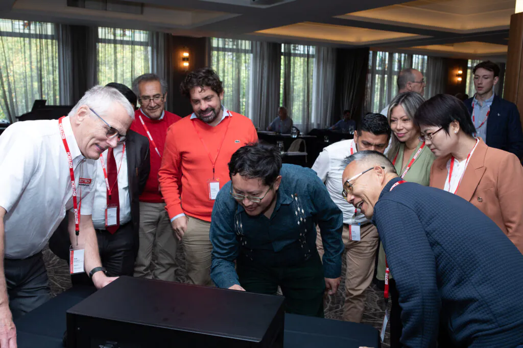 Paul Bramley and a group of international visitors gathered around a microK² on a table, seen from the rear, at an Isotech seminar, everyone leaning in to look at it.