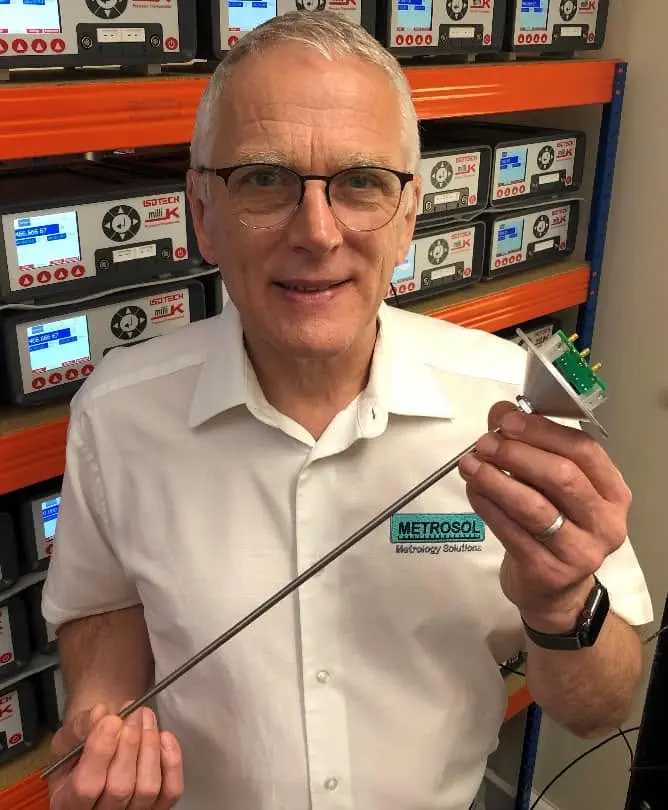 Paul Bramley, wearing a white Metrosol polo shirt, holding a circular pre-amplifier circuit board and a long probe, standing in front of a rack of Isotech microK and milliK instruments.